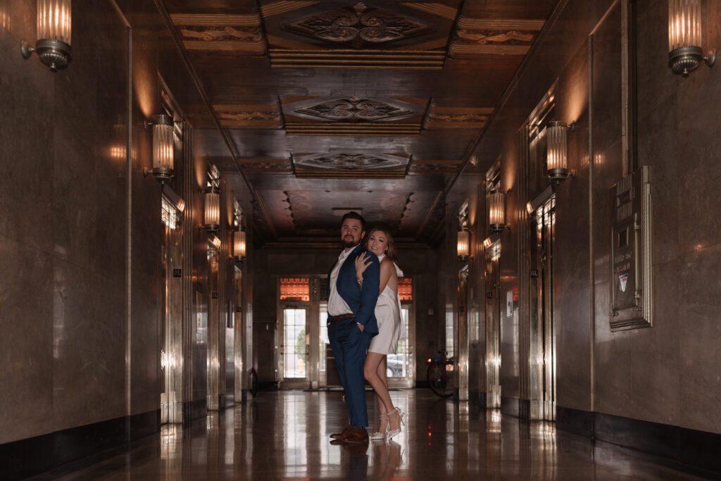 Engaged couple posing in the grand hallway of The Cardinal Hotel in Winston Salem, surrounded by art deco lighting and rich architectural details