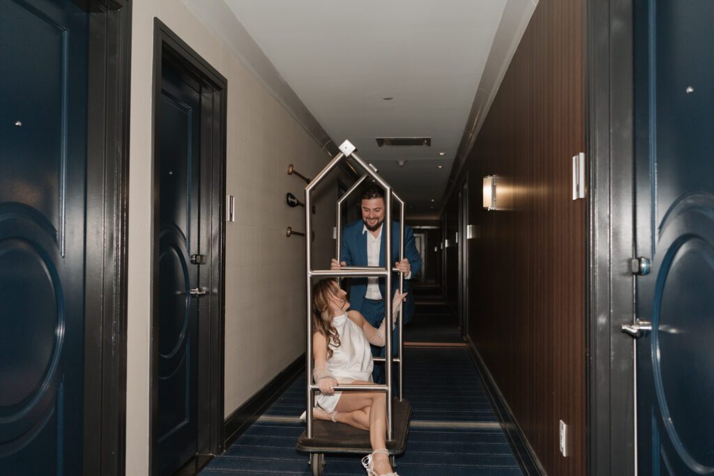 Engaged couple laughing as the groom pushes the bride and their dog on a luggage cart down a hotel hallway at The Cardinal Hotel in Winston Salem, NC