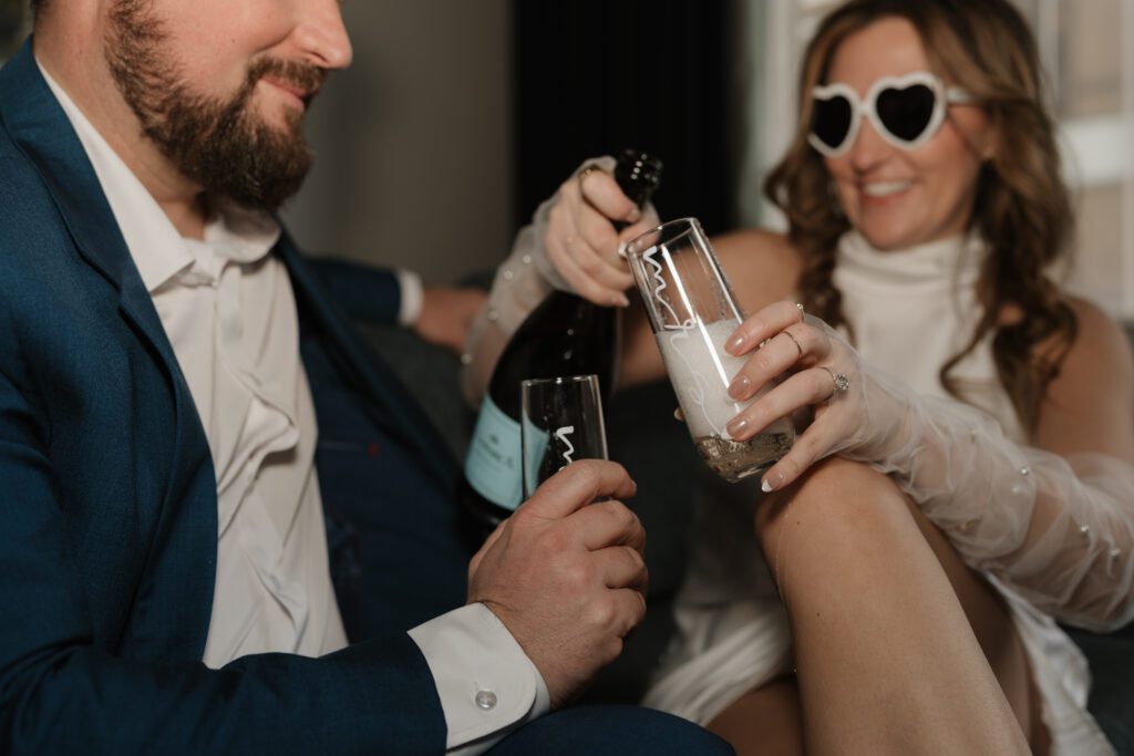 Bride-to-be wearing heart-shaped sunglasses and pearl gloves while pouring champagne during an engagement session at The Cardinal Hotel in Winston Salem, NC