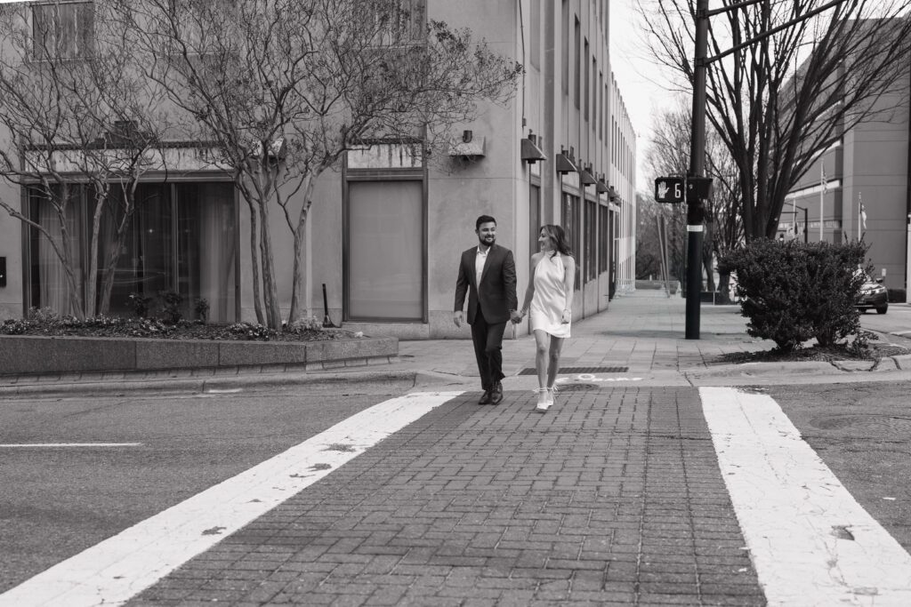 Engaged couple holding hands and smiling while walking across a crosswalk near The Cardinal Hotel in Winston Salem, NC